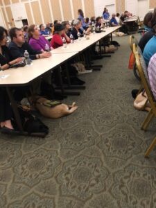 Guide dogs nap under long tables as their partners listen to a lecture.