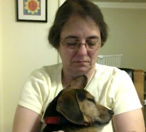 Small black and rust colored hound dog is sitting on a woman's lap with her head leaning up against her, eyes closed